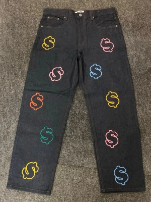 Svarta jeans med färgglada dollartryck - Unika svarta jeans från Fresh Flavours med färgglada dollar-tecken i rosa, blått, gult, grönt och orange. Rak passform och klassisk femficksmodell. Perfekta för dig som vill sticka ut med en kreativ och lekfull stil.
