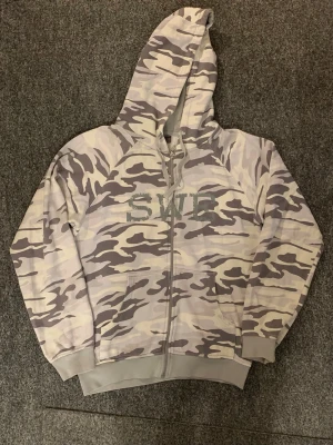 Grå camo hoodie med dragkedja SWE - Säljer en gråmelerad camo hoodie från SWE med dragkedja framtill och huva med dragsko. Hoodien har två fickor fram och ribbade muddar vid ärmar och nederkant. Perfekt för dig som gillar streetwear och vill sticka ut med ett snyggt camomönster.