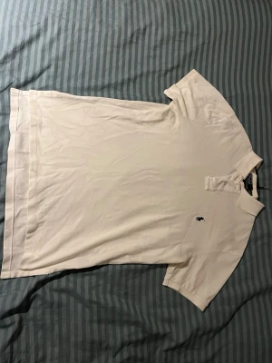 Vit klassisk pikétröja - Vit pikétröja med krage och korta ärmar. Tröjan har knappslå framtill och en liten broderad logga på bröstet. Perfekt till jeans eller shorts för en clean och enkel stil.
