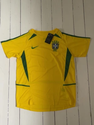 Retro Brasilien Nike fotbollströja - Säljer en gul Retro Brasilien fotbollströja från Nike med gröna detaljer och CBF-emblem på bröstet. Tröjan har korta ärmar och är tillverkad i ett lätt, ventilerande material med Dri-FIT-teknologi. Perfekt för träning eller match! Storlek L