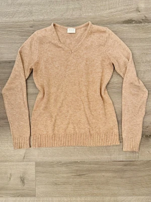 V-ringad beige tröja Vila XS - Mjuk och enkel beige  tröja från Vila med v-ringning och långa ärmar. Tröjan har ribbade muddar vid ärmslut och nederkant, vilket ger en snygg och avslappnad look. Perfekt att matcha med jeans eller kjol.
