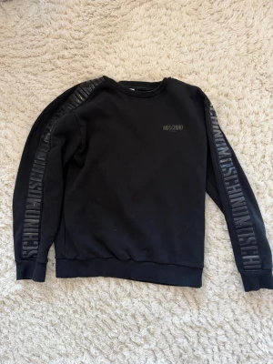 Svart Moschino sweatshirt - Äkta svart sweatshirt från Moschino med rund halsringning och ribbade muddar. Snygga detaljer med Moschino-logga tryckt längs båda ärmarna och liten logga på bröstet. Perfekt för en trendig och avslappnad stil. Ny pris ca 2800kr sälker för 799kr men hör av dig om du vill diskutera pris!