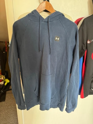 Blå hoodie från Under Armour - Mörkblå hoodie från Under Armour med klassisk logga på bröstet. Hoodien har snörning i huvan, stor magficka och långa ärmar. Perfekt för dig som gillar en enkel och sportig stil.