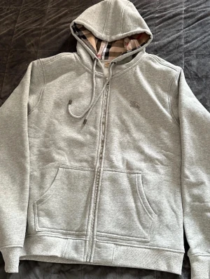 Grå zip hoodie från Burberry - Snygg grå zip hoodie från Burberry med klassiskt rutigt foder i huvan och diskret logga på bröstet. Hoodien har dragsko i huvan, två stora fickor fram och ribbade muddar vid ärmar och nederkant. Perfekt för en chill och stilren look.