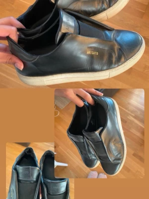 Svarta sneakers från Axel Arigato - Stilrena svarta sneakers i läder från Axel Arigato med vit sula och guldfärgad logga på sidan. Skorna har elastiska band över vristen istället för snören och en klassisk låg siluett. Perfekta för en clean och modern look. Defekt på insidan bak i skon, inget som syns utåt och inget som stör. 