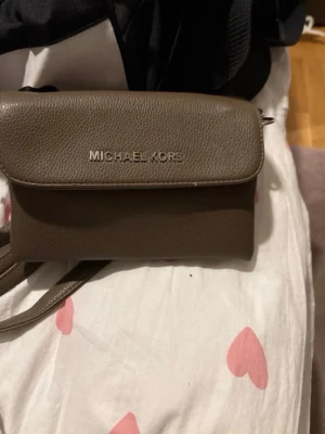 Brun axelväska från Michael Kors - Snygg brun axelväska från Michael Kors i slätt läder med silverfärgad logga på framsidan. Väskan har ett stilrent och kompakt format med justerbar axelrem, perfekt för att bära det viktigaste. Enkel design som funkar till många outfits.