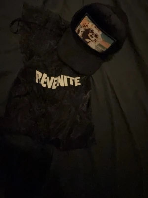 Svart keps från Revenite med tryck - Svart keps från Revenite med mesh på baksidan och ett färgglatt tryck framtill som föreställer en tecknad figur. Kepsen har böjd skärm och justerbar passform. Perfekt accessoar för en cool streetwear-look.