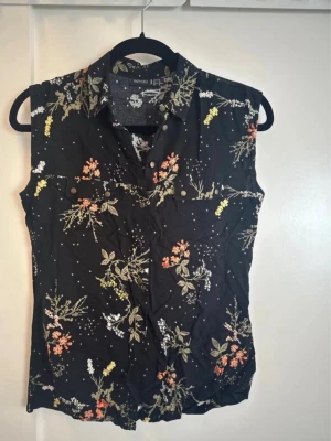 Svart blommig ärmlös blus Gina Tricot - Svart ärmlös blus från Gina Tricot med färgglatt blommönster i orange, vitt och gult. Blusen har krage, knäppning framtill och två bröstfickor med knapp. Perfekt för en trendig och avslappnad look.