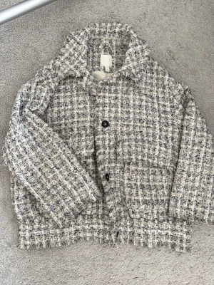 Tweed cardiganjacka från H&M - Säljer en snygg tweed cardiganjacka från H&M i beige, svart och vitt. Jackan har två stora fickor framtill, knappar och en boxy passform. Materialet är grovt vävt och ger en trendig, klassisk look. Perfekt för lager på lager under kyliga dagar.