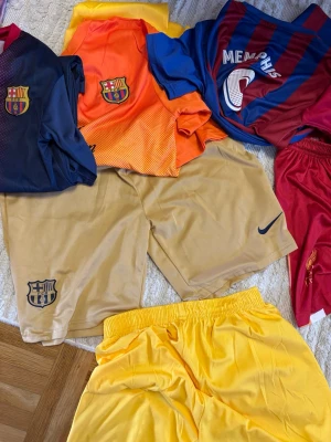 FC Barcelona matchställ och shorts - Flera FC Barcelona matchtröjor och shorts i olika färger: marinblå, orange, rödblå, guld och gul. Tröjorna har klubbmärke och vissa har spelarnamn, shortsen har klubb- och Nike-logga. Perfekt för fotbollsträning eller support.  Olika storlekar från S till M  och vill sälja dom för jag har inte på mig det mer. 