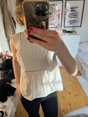 Vit ärmlös blus med volanger - Söt vit blus med volangdetaljer längs axlar och framsida. Blusen är ärmlös och har fina veckade detaljer framtill samt spetskant på volangerna. Perfekt för en fräsch och somrig look. Materialet känns lätt och luftigt.