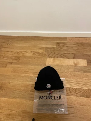 Svart Moncler mössa  - Svart ribbstickad mössa från Moncler med klassisk uppvikt kant och broderad logotyp framtill. Tillverkad i mjuk ull som håller dig varm och stilren under kalla dagar. Perfekt accessoar för en trendig vinterlook.