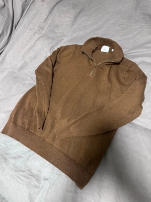 Brun stickad halvzip-tröja från Bläck - Snygg brun stickad tröja från Bläck med halv dragkedja och hög krage. Tröjan har ribbade muddar vid ärmslut och nederkant, perfekt för lager-på-lager. Enkel och stilren design som passar till det mesta.