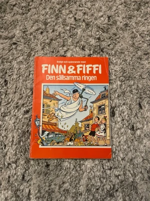 Finn & Fiffi: Den sällsamma ringen - Säljer seriealbumet 'Finn & Fiffi: Den sällsamma ringen' med färgglad och tecknad framsida. Boken har ett rött omslag med illustrationer av Finn och Fiffi i ett äventyr. Perfekt för dig som gillar roliga och spännande berättelser i serieformat.