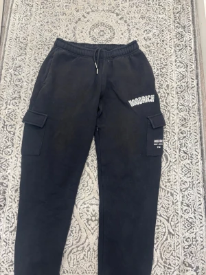 Svarta cargo mjukisbyxor Hoodrich - Säljer ett par svarta mjukisbyxor från Hoodrich med cargo-fickor på sidorna och tryckt logga på ena benet. Byxorna har resår i midjan med dragsko och är tillverkade i mjukt material som passar perfekt för chill eller streetwear.
