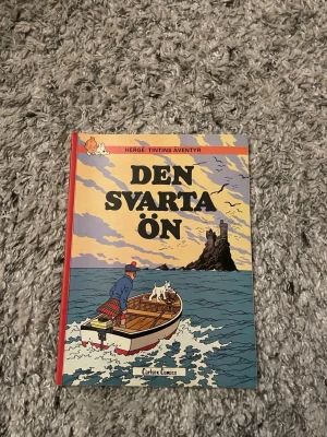 Tintins äventyr: Den svarta ön - Säljer seriealbumet 'Den svarta ön' från Tintins äventyr av Hergé. Omslaget är färgglatt med motiv av Tintin och Milou i en båt på havet mot en dramatisk ö. Boken har hårda pärmar och klassisk serietidningsstil.