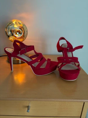 Röda höga klack-sandaler med remmar - Snygga röda sandaler med höga klackar och flera remmar över foten. Skorna har en justerbar spänne vid ankeln och en platåsula framtill. Materialet ser ut att vara mockaimitation och sulan är ljus. Perfekta för att lyfta vilken outfit som helst.