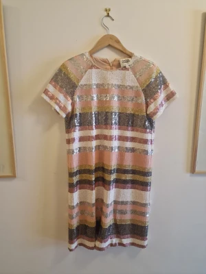 Randig paljettklänning Twist & Tango - Glittrig kortklänning från Twist & Tango med färgglada ränder i rosa, guld, silver och vitt. Klänningen har korta ärmar, rund halsringning och är täckt av paljetter för en extra festlig look. Perfekt för dig som vill sticka ut.