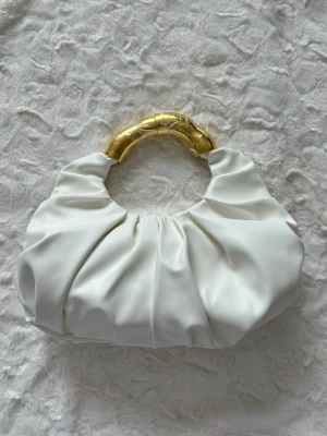 Vit handväska med guldfärgat handtag - Trendig vit handväska i mjukt, veckat material med ett rundat handtag i guldfärgad metall som ger en lyxig känsla. Väskan har en unik form och passar perfekt för dig som vill sticka ut med din stil.