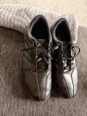 Svarta Adidas golfsneakers med klassiska ränder - Svarta sneakers från Adidas med de ikoniska tre ränderna på sidan. Skorna har lågt skaft, snörning och är tillverkade i en kombination av läder och syntetmaterial. Perfekta för en stilren och sportig look.
