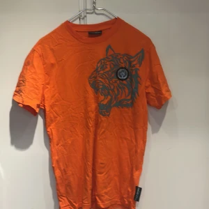 Orange t-shirt med tigertryck Kenzo - Cool orange t-shirt från Kenzo med stort tigertryck i grått på bröstet. Klassisk rund hals och korta ärmar. Perfekt för dig som vill sticka ut med en färgstark och trendig look.