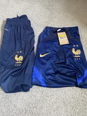Frankrike Nike träningsset blå/guld - Snyggt träningsset från Nike och franska landslaget, FFF. Setet består av en långärmad tröja och byxor i mörkblått med guldiga detaljer och emblem. Tröjan har halv dragkedja och mönster i blått. Perfekt för fotbollsträning eller sport.