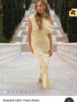 Ljusgul draperad satinklänning - Elegant långklänning i ljusgul satin med draperad design och oneshoulder-skärning. Klänningen har en figurnära passform och ett mjukt, glansigt material som ger en lyxig känsla. 