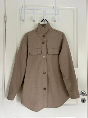 Beige kappa med knappar  -  beige kappa med knappar framtill och två bröstfickor. Kappan har en rak passform och hög krage. Från MSCH i storlek xs/s