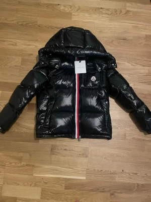 Moncler jacka Montbeliard - Snygg svart pufferjacka från Moncler med glansig finish, stor huva och klassisk logotyp på bröstet. Jackan har en rödvit dragkedja framtill och praktiska fickor. Perfekt för kalla vinterdagar och riktigt trendig stil.
