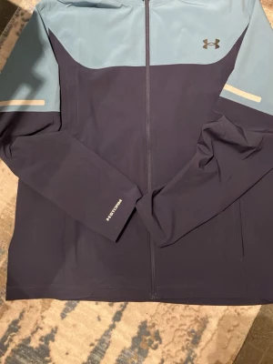 Blå vindjacka från Under Armour - Sportig vindjacka från Under Armour i mörkblått och ljusblått. Jackan har hel dragkedja framtill, långa ärmar och reflekterande detaljer på ärmarna. Perfekt för utomhusaktiviteter och träning.