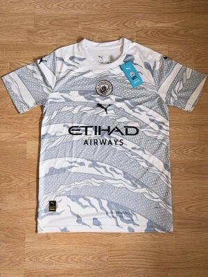 Manchester City bortatröja Puma - Snygg Manchester City bortatröja från Puma i vitt och ljusgrått med grafiskt mönster. Tröjan har klubbmärke på bröstet, svart Puma-logga och Etihad Airways-tryck. Tillverkad i lätt och ventilerande material, perfekt för fotbollsträning eller match.