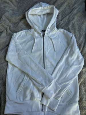 Vit hoodie med dragkedja från 157 - Vit hoodie från 157 med dragkedja framtill och huva med snören. Tillverkad i mjuk bomullsmix, perfekt för en chill och avslappnad stil. Två fickor framtill och ribbade muddar vid ärmslut och nederkant.