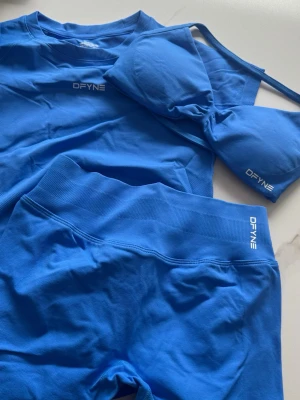 Blått träningsset från DFYNE - Pris kan diskuteras, ge förslag. Säljer ett blått träningsset från DFYNE som består av en t-shirt, sport-bh och shorts. Setet är tillverkat i ett stretchigt och mjukt material som passar perfekt för gym och träning. Alla delar har logotypen tryckt framtill och är designade för att sitta bekvämt.