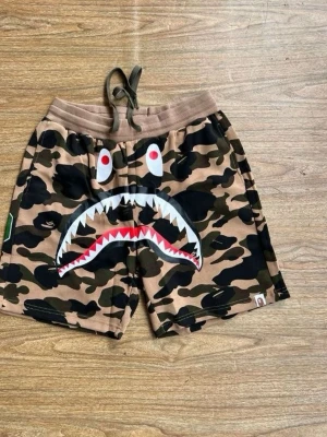 BAPE camo shorts med hajmotiv - Coola shorts från BAPE med klassiskt camouflage-mönster och hajmotiv på framsidan. De har snörning i midjan och är tillverkade i mjuk bomull. Perfekta för dig som vill sticka ut med streetwear-stil.