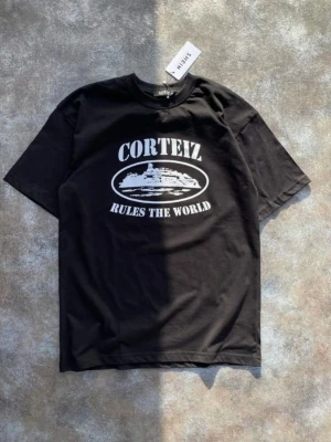 Svart Corteiz t-shirt med tryck - Svart t-shirt från Corteiz med stort vitt tryck på bröstet där det står 'CORTEIZ RULES THE WORLD' och en grafisk illustration. Klassisk passform och rund halsringning. Perfekt för dig som gillar streetwear och vill sticka ut.
