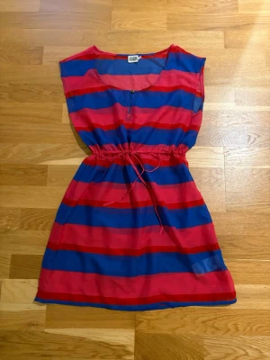 Randig strandklänning från Twist & Tango - Färgglad strandklänning H&M breda blå och röda ränder. Modellen är ärmlös, har rund halsringning och snörning i midjan för en avslappnad passform. Tillverkad i ett lätt syntetmaterial som passar perfekt för varma sommardagar. Längd 89 cm