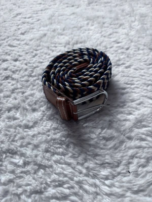 Randigt flätat bälte med läderdetaljer - Snyggt flätat bälte med randigt mönster i marinblått, vitt och brunt. Bältet har detaljer i brunt skinn och ett klassiskt metallspänne. Perfekt accessoar för att lyfta din outfit med en cool touch.