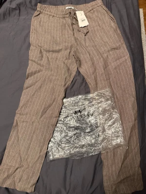 Beige linnebyxor från John Henric - Snygga beige linnebyxor från John Henric med vita tunna vertikala ränder. Byxorna har dragsko i midjan och raka ben, vilket ger en avslappnad och trendig look. Perfekta för dig som gillar stilrena och luftiga plagg i naturligt material. Helt nya säljer då jag fått fel storlek