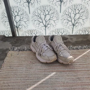 Vita Adidas Falcon sneakers - Säljer ett par vita Adidas Falcon sneakers med chunky sula och meshdetaljer. Skorna har klassisk Adidas-logga på plösen och sidorna, samt grå och beige inslag. Perfekta för dig som gillar retrostil och streetwear.