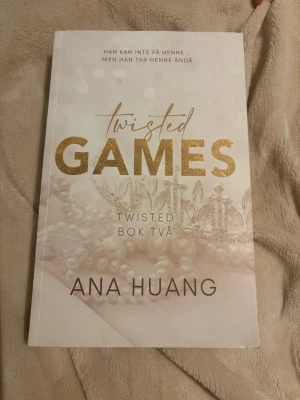 Twisted Games av Ana Huang - Twisted Games är andra boken i Twisted-serien av Ana Huang. Omslaget är vitt med guldiga detaljer och en elegant krona. Boken är en romantisk berättelse fylld av känslostormar, maktspel och förbjuden kärlek. Perfekt för dig som gillar spännande och dramatiska romaner.