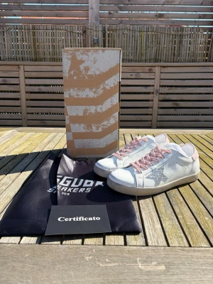 Vita Golden Goose sneakers med glitterstjärna - Snygga vita sneakers från Golden Goose med rosa skosnören och häl, samt en glittrig silverstjärna på sidan. Skorna har en klassisk låg siluett och är tillverkade i läder med detaljer i mocka. Perfekta för dig som vill ha en unik och trendig look.
