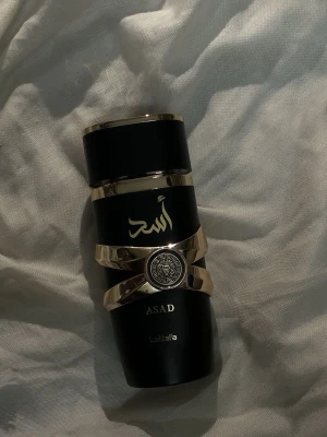 Lattafa Asad parfym svart/guld - Snygg parfymflaska från Lattafa i svart med guldiga detaljer och arabiska tecken. Flaskan har en modern, lyxig design med metalliska inslag och en dekorativ emblemring. Perfekt för dig som gillar exklusiva dofter och stilren look.