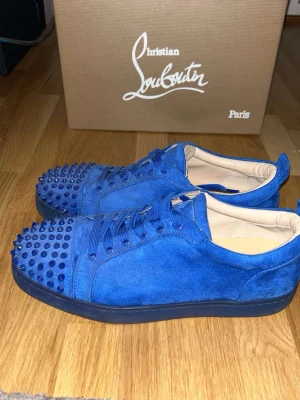 Blåa Christian Louboutin sneakers - Snygga blåa sneakers från Christian Louboutin i mocka med matchande blå spikar där fram och klassisk röd sula. Perfekta för dig som vill sticka ut med exklusiva detaljer och unik design. En spik har lossnat men man får med 5 extra spikar!!🫡😊