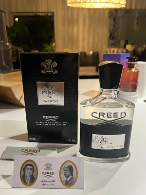 Creed Aventus Eau de Parfum 100ml - Creed Aventus är en exklusiv parfym i en elegant, genomskinlig glasflaska med svart och silverdetaljer. Flaskan rymmer 100 ml och har en svart kork med silveremblem. Doften är känd för sin fräscha och sofistikerade karaktär.