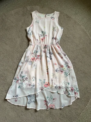 Vacker ljusrosa blommig klänning - Söt ljusrosa klänning med blommigt mönster och omlottliknande nederdel på framsidan. Ärmlös modell med resår i midjan, rund halsringning och knapp baktill vid nacken! Perfekt för varma dagar och har en luftig känsla🌸Klänningen har en underkjol så den inte är genomskinlig och meshtyg i blommigt utanpå❣️Passar så bra till skolavslutning, events, firanden, födelsedag, en varm kväll, midsommar, semester eller när man bara vill vara fin💕Storlek 152, passar tonåringar! Använd några gånger, inge fel❤️