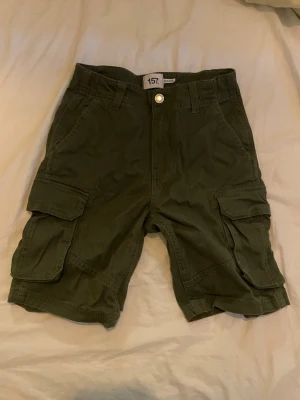 Olivgröna cargoshorts från 157 - Olivgröna cargoshorts från 157 med flera stora fickor på sidorna och klassisk knappstängning framtill. Shortsen har en avslappnad passform och är perfekta för varma dagar. Tillverkade i bomull för en skön känsla.