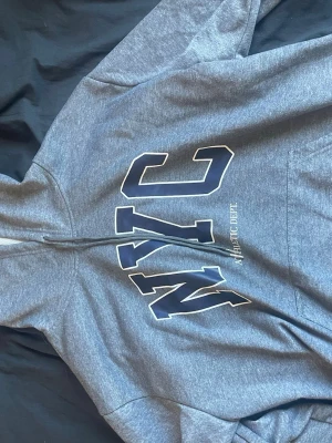 Grå hoodie med NYC-tryck - Snygg gråmelerad hoodie med stor NYC-text i vitt och mörkblått på framsidan samt texten 'ATHLETIC DEPT' under. Klassisk huva och känguruficka framtill. Perfekt för en avslappnad streetwear-look. Mjuk och bekväm bomullsblandning. Kontakta gärna ❤️