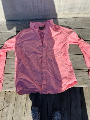 Rosa skjorta från Polo Ralph Lauren - Klassisk rosa skjorta från Polo Ralph Lauren med broderad logga på bröstet. Skjortan har lång ärm, knappar framtill och en snygg krage. Perfekt för en stilren och avslappnad look.