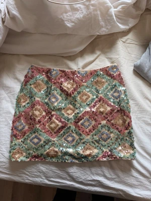 Paljettkjol med färgglatt mönster - Snygg kortkjol täckt av paljetter i pastelliga färger som rosa, guld, blått och grönt. Kjolen har ett geometriskt rutmönster och är perfekt för dig som vill sticka ut. Ingen synlig dragkedja eller knappar, bara massor av glittrande vibes.