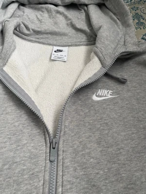 Grå zip hoodie från Nike - Snygg gråmelerad hoodie från Nike med dragkedja och vit broderad logga på bröstet. Utrustad med huva och dragsnören samt mjukt, vitt fleecefoder på insidan. Perfekt för chill dagar eller när du vill ha en sportig look. Tror det är s/xs